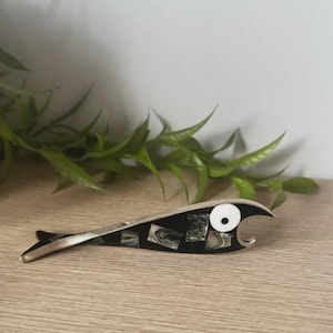 May include: A fish-shaped bottle opener crafted from silver-toned metal, featuring a black body inlaid with iridescent abalone shell segments. The design includes a prominent white circular eye with a black center, creating a stylized fish aesthetic.