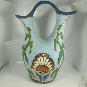 Puede incluir: Un jarrón de cerámica azul claro con un asa única y una textura moteada. El jarrón presenta diseños de hojas verdes y doradas, y un patrón de sol con formas geométricas. El borde y el asa son azules con ribete dorado.