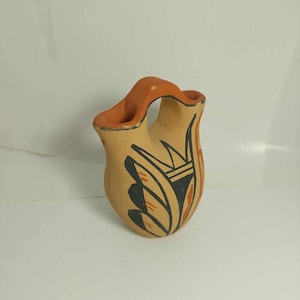 May include: A small, tan-coloured ceramic vessel featuring a bridge-style handle connecting two openings. The exterior is decorated with hand-painted black and orange geometric patterns, including leaf-like shapes and lines on a matte clay surface.