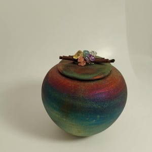 May include: A spherical, iridescent ceramic jar with a lid, displaying a range of colours from blue to red. The lid is decorated with a cluster of colourful gemstones and a small, brown stick. The jar is likely for storage or decorative use.
