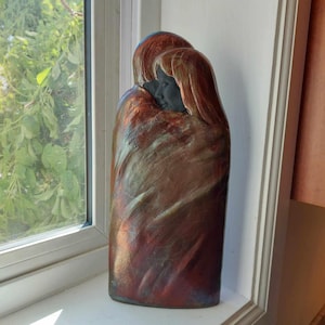 Könnte beinhalten: Eine Keramikskulptur von zwei sich umarmenden Figuren mit einem wirbelnden, abstrakten Design in Braun-, Gold- und Grüntönen.