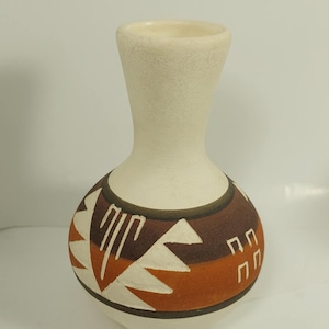 Peut inclure: Un vase en céramique de couleur crème avec une base bulbeuse et un col fin. La base présente un motif géométrique marron, orange et blanc. Le vase a une finition lisse et mate et une petite ouverture en haut.