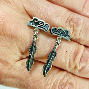 May include: Silver earrings with an eagle head design and a dangling feather pendant. The earrings have etched details, reflecting a Native American aesthetic. The feather pendants add a touch of movement and detail.