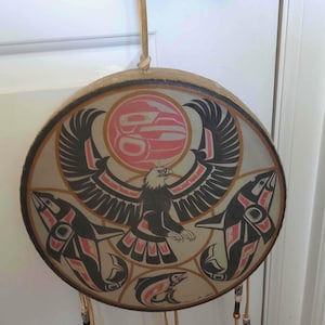 May include: A circular drum featuring a painted design of a central eagle with wings spread, positioned below a pink circular sun. Two orcas flank the eagle, with a smaller fish at the bottom. The drum is suspended by a light-colored leather cord.