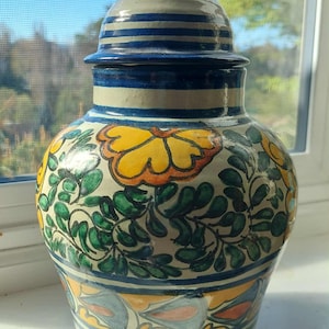 Puede incluir: Un jarrón de cerámica con tapa, con un diseño pintado a mano. El jarrón tiene una base color crema con rayas azules y está adornado con motivos de mariposas y flores amarillas. La tapa también es crema con rayas azules. El jarrón mide unos 30 cm de alto.