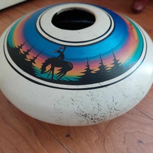 Könnte beinhalten: Eine weiße, runde Keramikvase mit einer breiten Öffnung. Die Vase hat ein farbenfrohes, gemaltes Design eines Regenbogens, Bäumen und einer Hirschsilhouette. Die Vase hat eine abgenutzte, rissige Oberfläche.