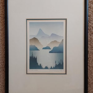 May include: Framed landscape art print depicting a tranquil mountain and lake scene in shades of blue, grey, and brown. The artwork is matted and framed in a black frame. The print includes the text "Ruby Lake" and a signature.