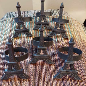 Peut inclure: Un ensemble de six ronds de serviette en métal brun foncé, chacun présentant une miniature détaillée et tridimensionnelle de la tour Eiffel fixée sur un anneau circulaire. Les structures ont une finition texturée et rustique.
