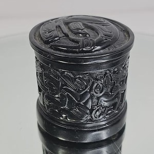 May include: A black, cylindrical container with a lid, featuring detailed carvings of figures and designs. The container's surface is highly detailed, with the carvings creating a textured appearance. The lid also has a carved design. The container is displayed on a reflective surface.