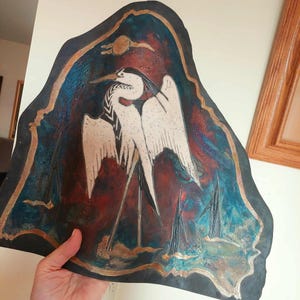 Könnte beinhalten: Ein unregelmäßig geformtes Kunstwerk mit einem Reiher-Design. Das Kunstwerk hat einen dunklen Rand mit goldenen Akzenten. Der Hintergrund wechselt von Rot zu Blau, mit einem weißen Reiher in der Mitte. Der Reiher ist in Schwarz und Weiß detailliert.