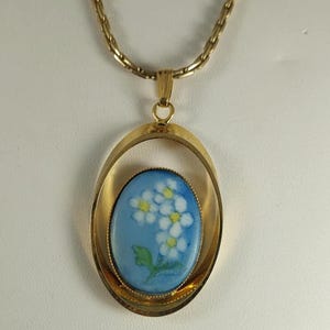 May include: Gold-toned pendant necklace featuring an oval-shaped pendant with a light blue background and a floral design. The flowers are white with yellow centers and green leaves. The necklace has a delicate chain.