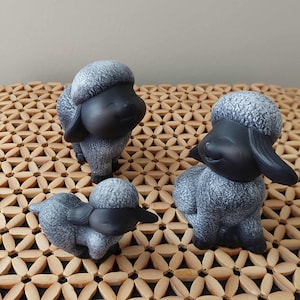 May include: Three small sheep figurines with smooth black faces and textured grey wool bodies. Each sheep has a smiling expression with closed eyes, long drooping ears, and a rounded head. The figurines are arranged in a group on a flat surface.