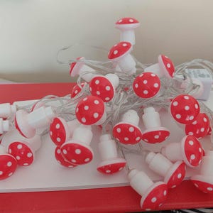 Peut inclure: Une guirlande lumineuse décorative avec des ampoules en forme de champignon. Chaque champignon a un chapeau rouge à pois blancs et une base blanche. Les lumières sont enfilées sur un fil transparent, créant un aspect fantaisiste et festif.