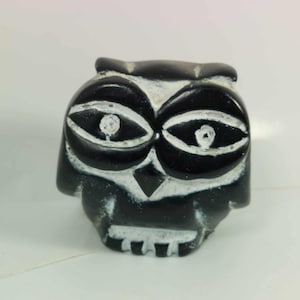 May include: A carved black owl figurine with white detailing. The owl features large, stylized eyes and a detailed beak. The figurine is likely made of stone or a similar material, and it has a smooth, polished finish. The owl is a decorative item.