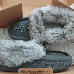 May include: A pair of grey suede moccasin slippers featuring a thick grey faux fur trim around the collar and a geometric beaded pattern on the top. A small brown rectangular card with the text "Win your next pair" is placed in the box.