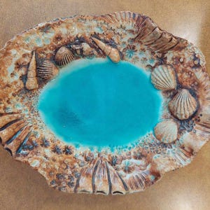 May include: An oval ceramic dish featuring a bright turquoise centre surrounded by a textured border of brown and tan sand-like patterns. The rim is decorated with various three-dimensional sculpted seashells, including spiral and fan-shaped shells.