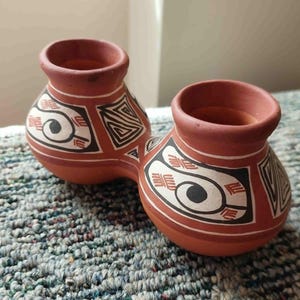 Op de afbeelding: Twee terracotta keramische vazen, verbonden aan de basis. Elke vaas heeft een brede opening en is versierd met zwarte, witte en rode geometrische patronen. De vazen staan op een gestructureerd, blauw en grijs oppervlak.