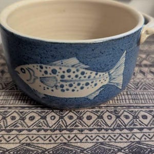 May include: A ceramic bowl with a blue exterior and a cream-coloured interior. The bowl features a white fish design on the side. The bowl has a handle and is sitting on patterned fabric.
