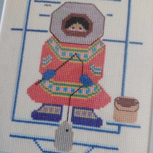 May include: Cross-stitch embroidery of a person wearing a red and blue patterned coat and a white hooded hat, holding a fishing rod with a fish on the line. The person is standing on a blue and white patterned surface with a brown bucket to the right.