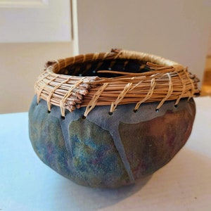 May include: A small, round, dark gray ceramic bowl with a woven rim of natural brown reeds. The bowl has a mottled, textured surface with hints of blue and purple.