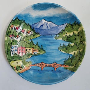 May include: Hand-painted ceramic plate featuring a scenic landscape. The plate depicts a blue river winding through green hills, with a snow-capped mountain in the background. A bridge spans the river, and colorful buildings dot the landscape. The plate's rim is a darker blue.