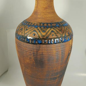 Könnte beinhalten: Eine hohe, braune Keramikvase mit strukturierter Oberfläche. Die Vase hat ein Band aus blauen und beigen geometrischen Mustern um den Hals. Die Vase hat einen schmalen Hals und einen breiteren Körper, geeignet für die Inneneinrichtung.