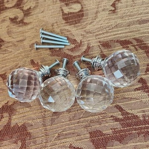 May include: Four clear, faceted crystal cabinet knobs with silver-toned hardware and a small pile of silver screws. The knobs are spherical and reflect light, creating a sparkling effect. Ideal for furniture upgrades.