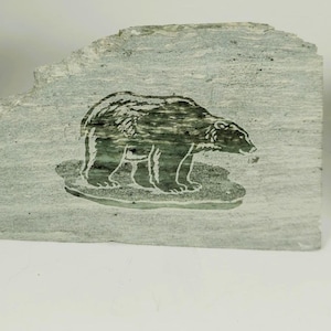 Peut inclure: Une pierre rectangulaire gris clair avec une image gravée d'un ours. L'ours est vert foncé avec des contours blancs, debout sur une base vert clair. La pierre a une surface rugueuse et texturée.