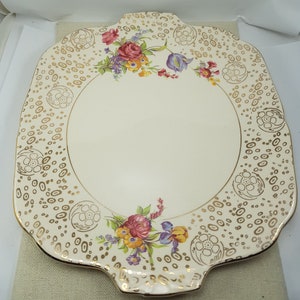 Puede incluir: Un plato para pastel de cerámica blanca con un borde festoneado y ribete dorado. El plato presenta un diseño floral con flores rosas, amarillas y moradas. El centro del plato es blanco liso.