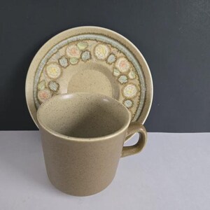 Könnte beinhalten: Ein gesprenkeltes, hellbraunes Keramik-Tassen- und Untertassen-Set. Die Tasse hat eine abgerundete Form mit einem Henkel. Die Untertasse hat ein dekoratives Blumenmuster in Pastellfarben. Das Set ist für Heißgetränke konzipiert.