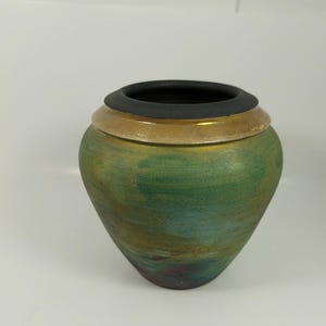 Könnte beinhalten: Eine handgefertigte Keramikvase mit rundem Körper und breiter Öffnung. Die Vase hat einen Farbverlauf aus Grün- und Blautönen, mit einem goldenen Band und einem schwarzen Rand. Die Keramik hat eine strukturierte Oberfläche.