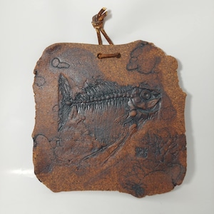 May include: A brown, rectangular ceramic wall hanging with a fish skeleton design. The fish skeleton is dark brown and detailed, set against a lighter brown background. A leather cord is attached for hanging.