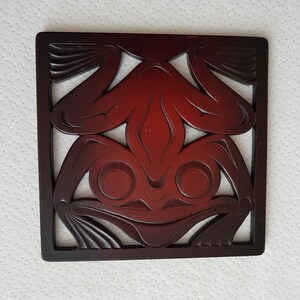 May include: A square, dark reddish-brown wooden coaster featuring a carved frog design. The frog is stylized with large, round eyes and a smiling mouth, set within a square frame. The coaster is likely for decorative use.