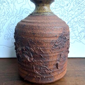 Puede incluir: Un jarrón de cerámica cilíndrico con un exterior rugoso, de color marrón oscuro y texturizado con estrías horizontales. El cuello del jarrón es liso, acampanado y tiene un acabado de esmalte moteado en verde oliva, contrastando con el cuerpo oscuro.
