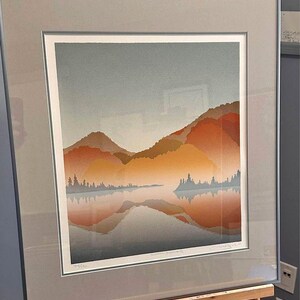 以下が含まれることがあります： 湖と背景の山を描いた風景画の額装されたプリント。山はオレンジ、赤、黄色の色合いで構成されています。湖は山を反射した明るい光を帯びた反射する青色です。プリントはアーティストによって署名されています。