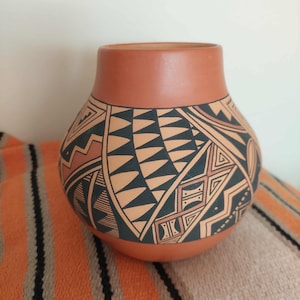May include: A terracotta-coloured clay pot with a rounded body and a wide opening. The exterior features intricate black geometric patterns, including triangles, zig-zag lines, and diamond shapes, painted directly onto the smooth matte surface.