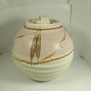 Può includere: Un vaso rotondo in ceramica con coperchio. Il vaso è principalmente rosa chiaro con pennellate marroni e una base color crema. Il coperchio ha una piccola maniglia. Il vaso ha un aspetto strutturato e fatto a mano.