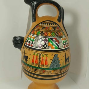 May include: A tan ceramic vessel with a black handle and two black animal figures. The vessel is adorned with colorful geometric patterns and images of llamas and a pyramid. The base is a pedestal shape.