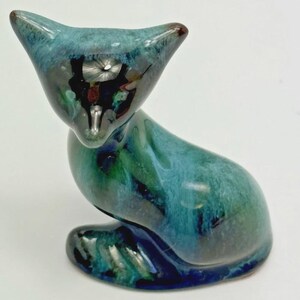 May include: A ceramic cat figurine featuring a glossy, mottled glaze in shades of deep teal, forest green, and dark blue. The cat is depicted in a seated position with its head tilted slightly downward, showcasing a smooth, reflective surface finish.