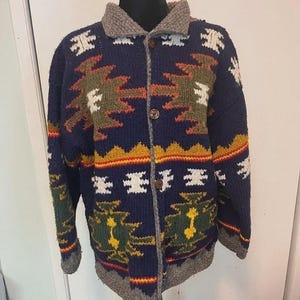 Puede incluir: Un cárdigan azul marino con botones y un estampado de inspiración suroeste. El suéter presenta un cuello y ribetes grises, con un diseño en tonos naranja, verde, amarillo y blanco. El suéter se exhibe en un maniquí.