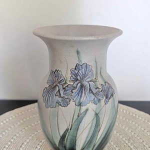 May include: A ceramic vase with a wide mouth and a light grey finish. The vase is decorated with blue iris flowers and green leaves. The vase is on a round, beaded placemat.