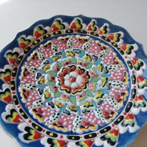 Puede incluir: Un plato decorativo de cerámica con un vibrante diseño floral. El plato presenta un borde azul con un borde festoneado y un patrón central de flores rosas, rojas, amarillas y verdes. El diámetro del plato es de aproximadamente 25 cm.