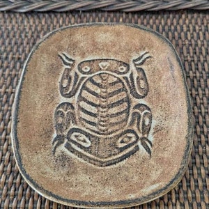 May include: A small, oval-shaped ceramic dish with a brown glaze. The dish features a raised, embossed design of a frog in the centre.