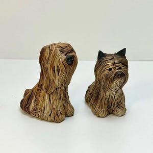 Op de afbeelding: Twee kleine keramische hondenbeeldjes. De hond links is bruin en wit met lang haar. De hond rechts is bruin met zwarte oren en lang haar.