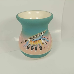 Peut inclure: Un petit vase en céramique de couleur turquoise avec un motif rose et blanc représentant une coiffe amérindienne avec des plumes.