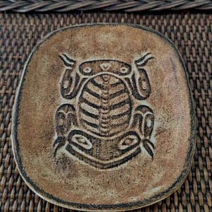 Peut inclure: Une assiette carrée en céramique marron aux coins arrondis. L'assiette présente un motif de grenouille stylisée en relief au centre. L'assiette a une surface texturée et est posée sur une surface tissée marron.