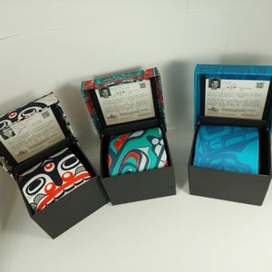 May include: Three neckties in black boxes. Each tie features a unique, colourful, abstract design. The boxes are open, revealing the ties and a printed card. The tie designs include red, black, white, and teal patterns.