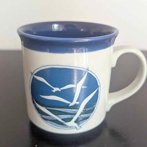 May include: A ceramic mug with a white exterior and a blue interior rim. The front features a circular graphic depicting white seagulls flying over blue and green waves. The mug has a curved handle on the right side and a smooth, glossy finish.