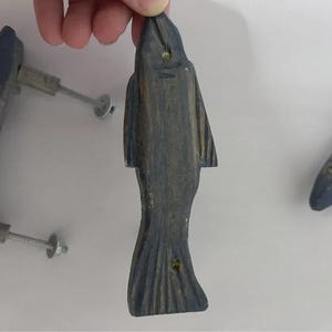 Pode incluir: Uma mão segura um puxador de armário em forma de peixe de madeira esculpida. O puxador é pintado em tons de cinza e azul, com um acabamento texturizado. O peixe tem um design detalhado de barbatana e cauda, e um parafuso de metal está preso.