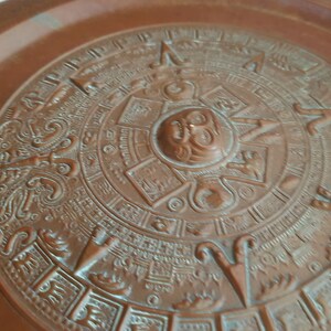 Puede incluir: Un plato de color cobre con un diseño en relieve detallado del calendario azteca. El calendario es circular y presenta un dios sol central con intrincados glifos y símbolos que lo rodean.